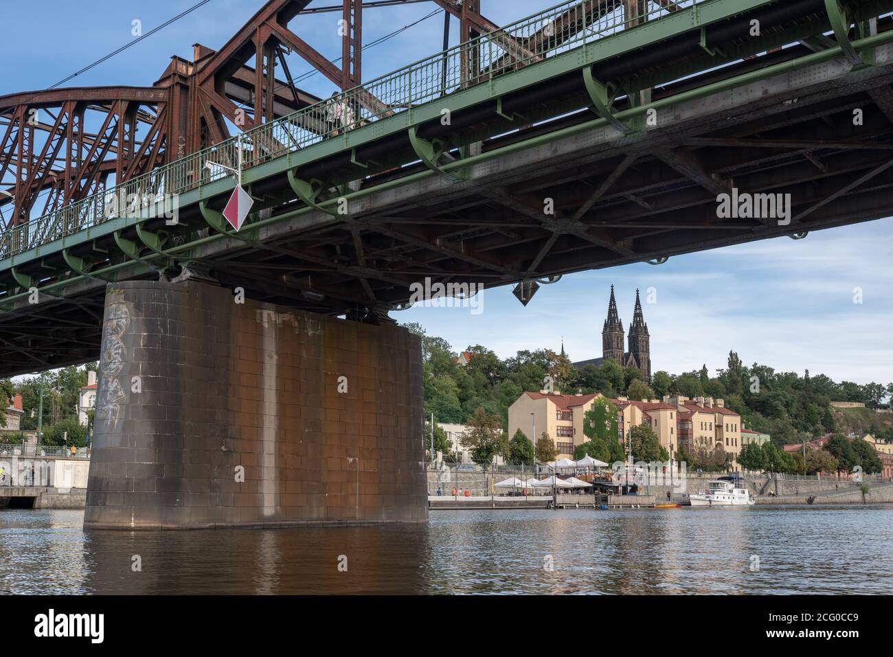 A view from under historic girder steel rail bridge over Vltava river in Prague towards Vysehrad castle and riverside embankment. Stock Photo