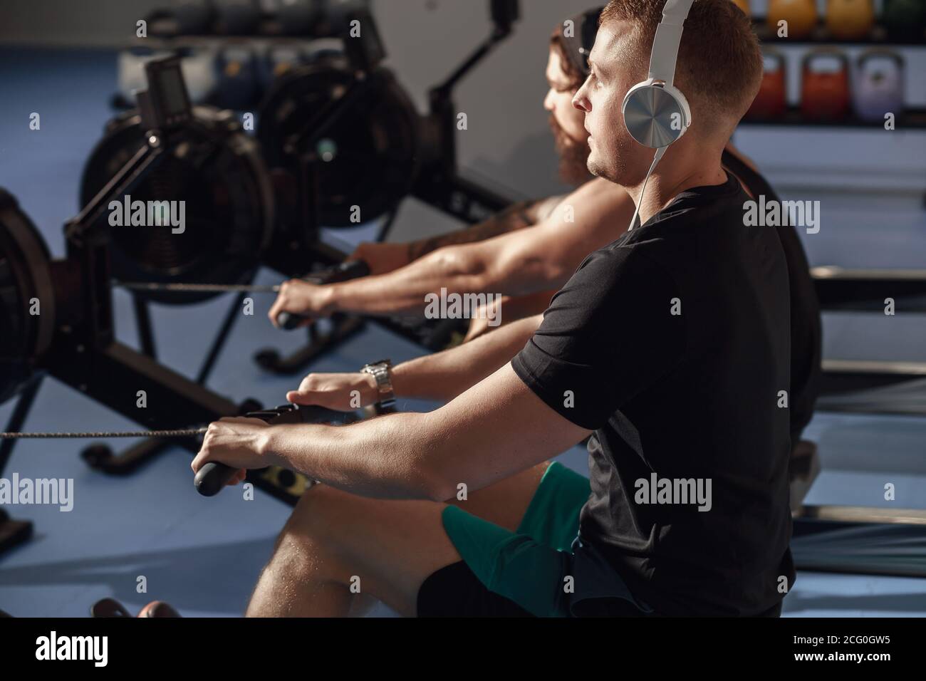 young sportsmen having hard workout on rowing machines in gym. Stock Photo