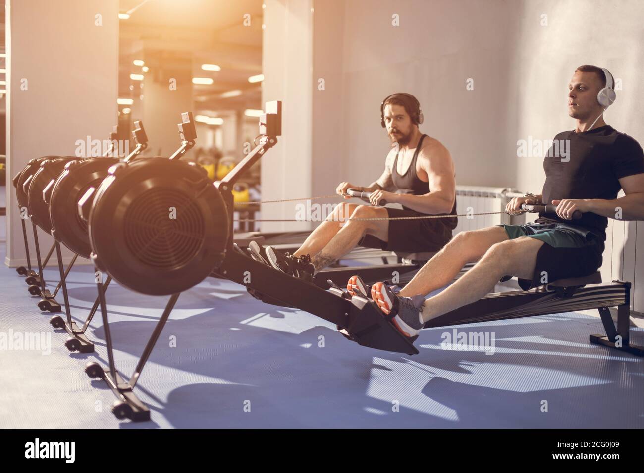 young sportsmen having hard workout on rowing machines in gym. Stock Photo