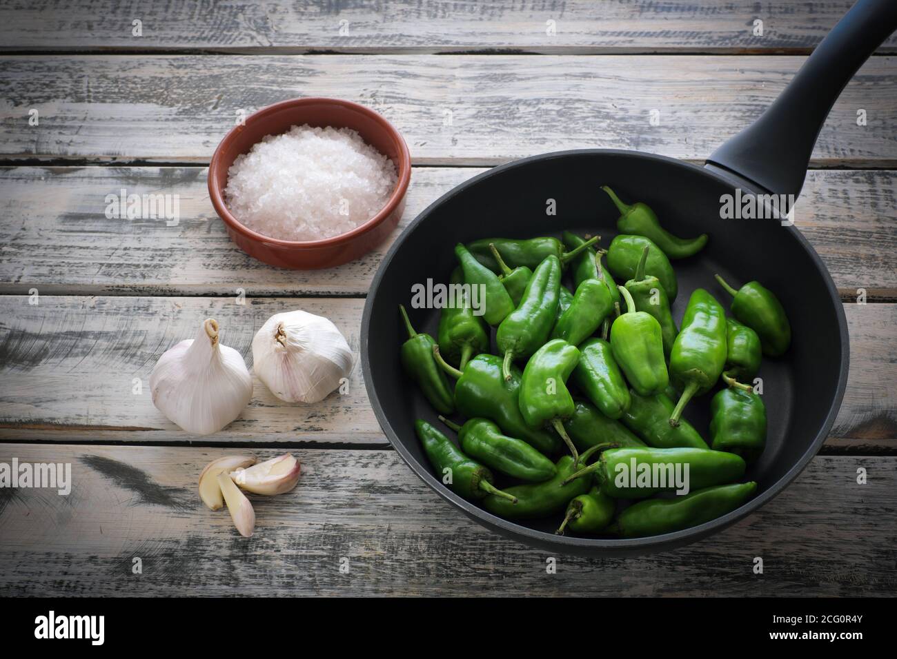 A black pan filled with raw pimentos, nicely arranged on a white shabby chic wooden plate, next to it a bowl of coarse sea salt and garlic Stock Photo