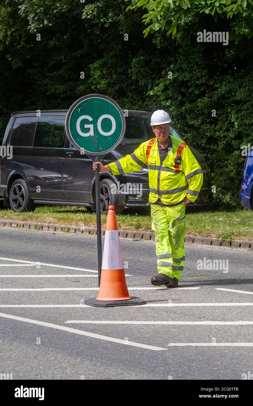 A traffic control worker in high visibility clothing with a traffic control lollipop with the green go side showing standing next to a cone in the roa Stock Photo