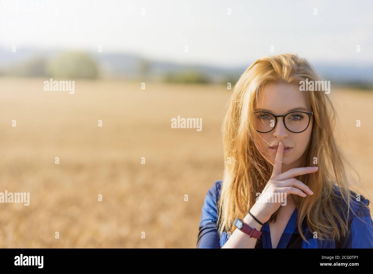 Portrait of young woman wearing glasses and a blue plaid shirt gesturing Shhh in corn field Horizontally. Stock Photo