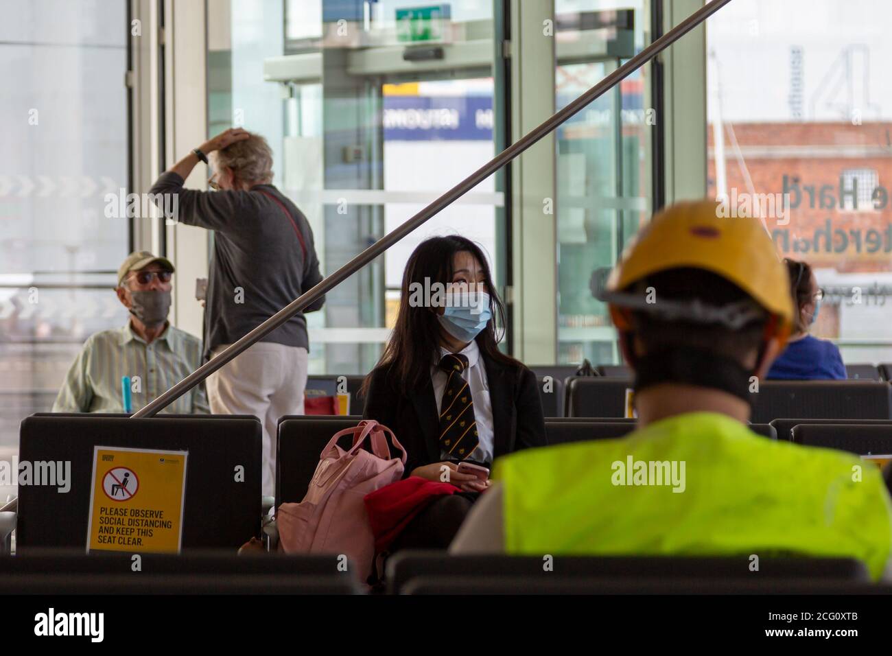 A young Asian school girl in uniform wearing a medical face mask or face covering will sitting waiting for a bus in a bus station Stock Photo