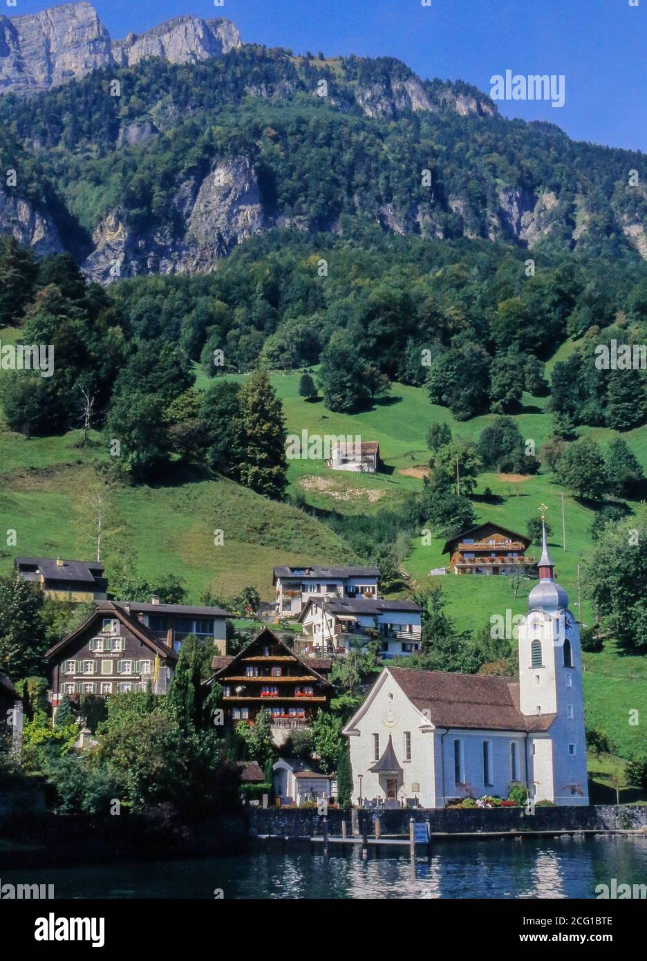 White village church of Saint Idda on shore of Lake Lucerne with mountain pastures and rocky outcrops in the background behind lakeside village Bauen, Stock Photo