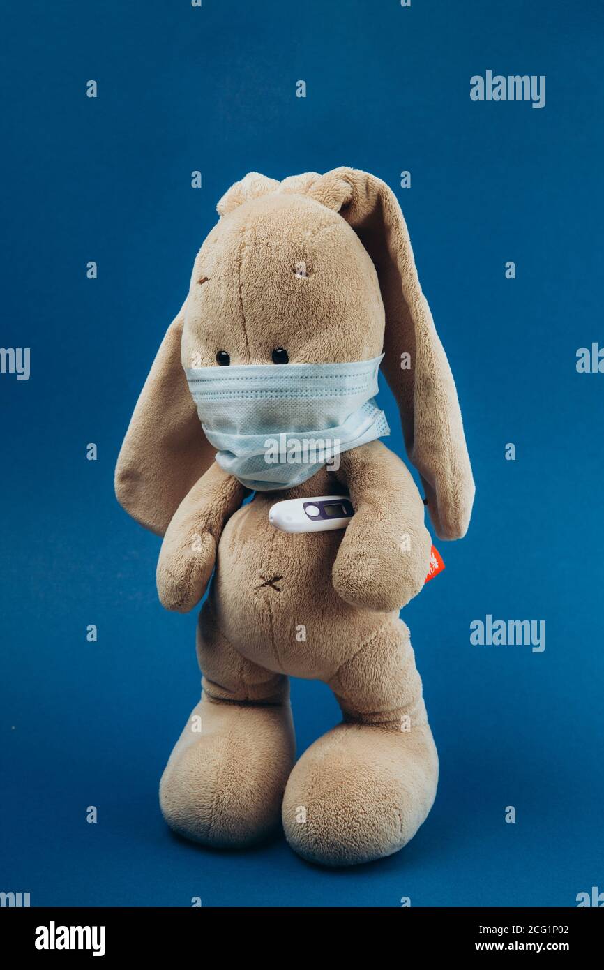 A toy bunny wears a protective medical mask with a thermometer under his paw on a blue background. Stop coronavirus. Medicine concept. Stock Photo