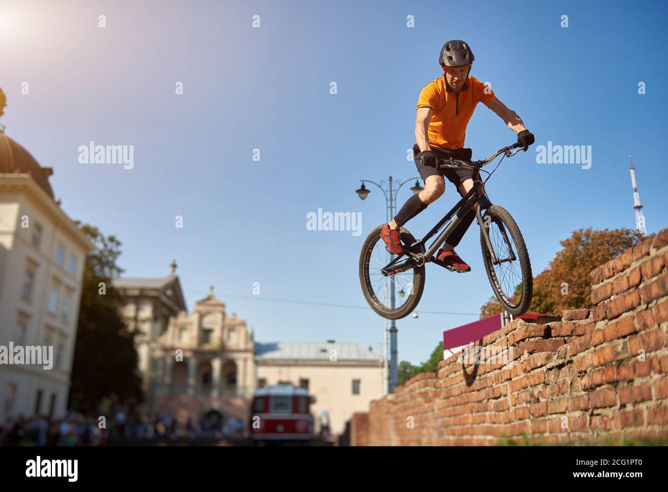Horizontal snapshot of a freestyle biker jumping from a brick wall making extreme stunt over blue sky, low angle view, urban buildings on background Stock Photo