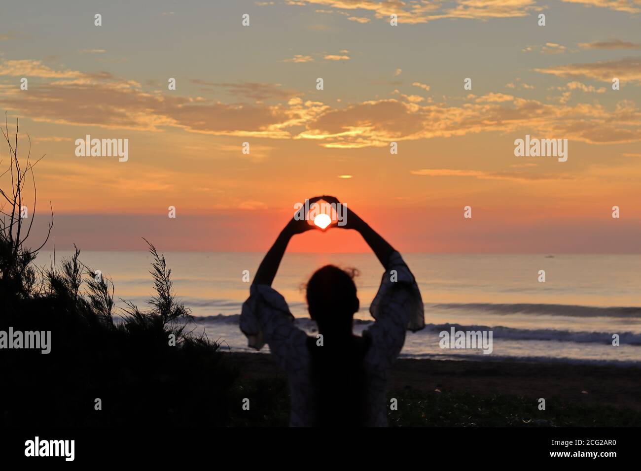 An Indian young woman, with the help of fingers on the top of her head keeping the bright sun in the center, makes a heart shape. sunrise at beach in Stock Photo