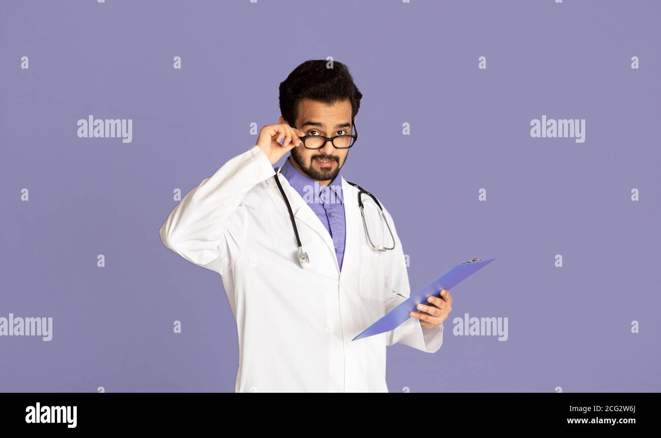 Young Indian doctor in uniform and glasses holding clipboard on violet background Stock Photo
