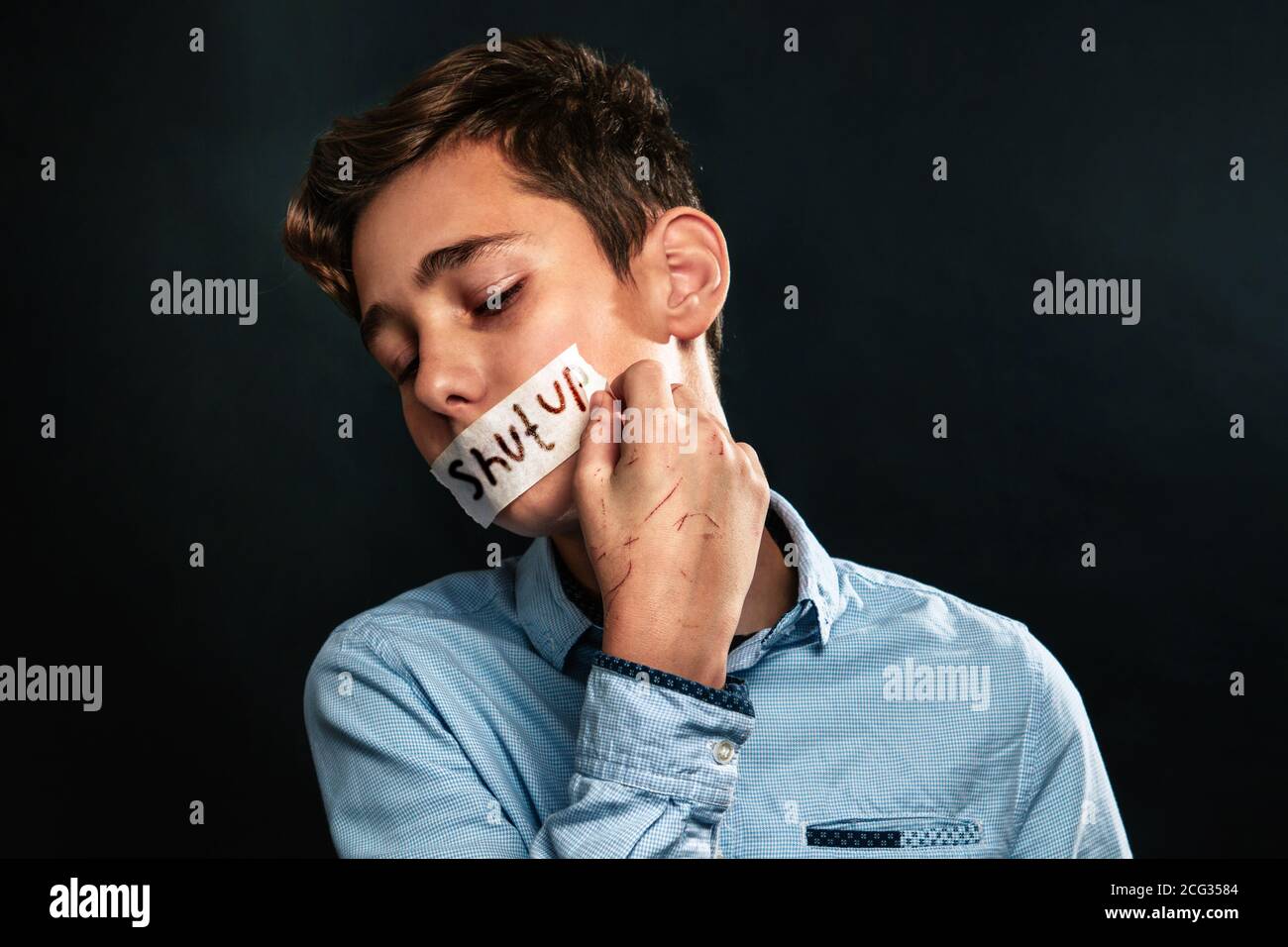 The concept of domestic violence. A teenage boy with his mouth taped shut rips off a tape with the words shut up with his scratched hands. Black backg Stock Photo