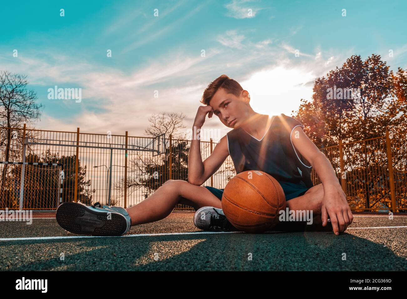 Sports and basketball. A young teenager in a blue tracksuit poses with a ball while sitting on a sports field. Blue sky in the background. Copy space. Stock Photo