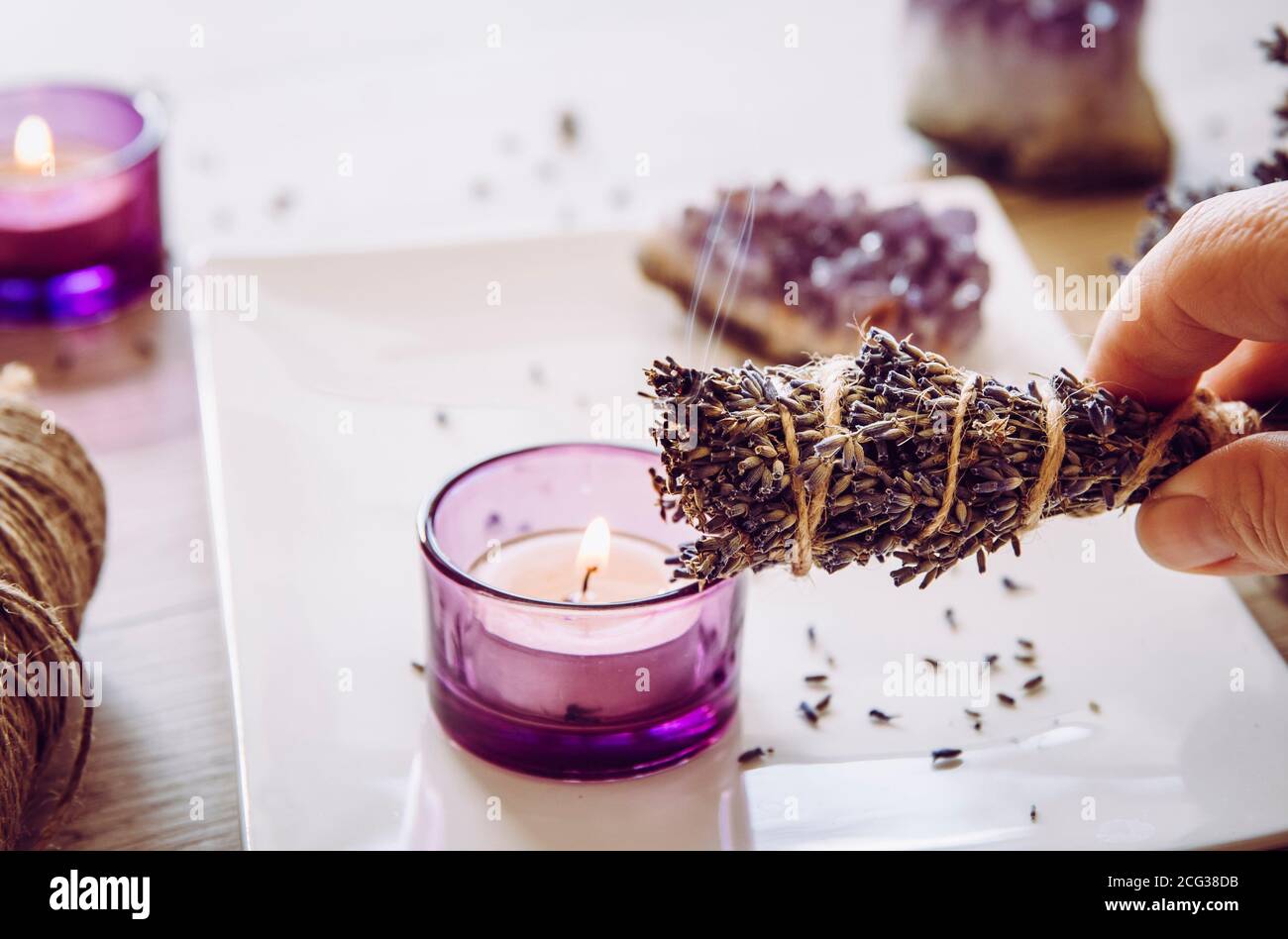 Person holding homemade herbal lavender (lavendula) smudge stick with smoke coming out, candles and amethyst crystal clusters for decoration. Stock Photo