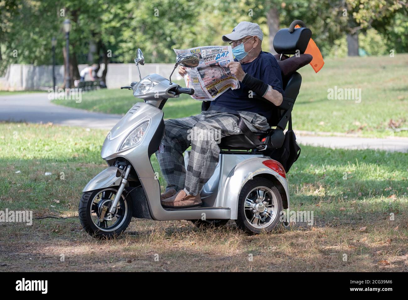 An older man wearing a mask over his mouth reads a foregn language newspaper on his 3 wheeled motor scooter in a snady spot in a park. In Queens, NYC. Stock Photo