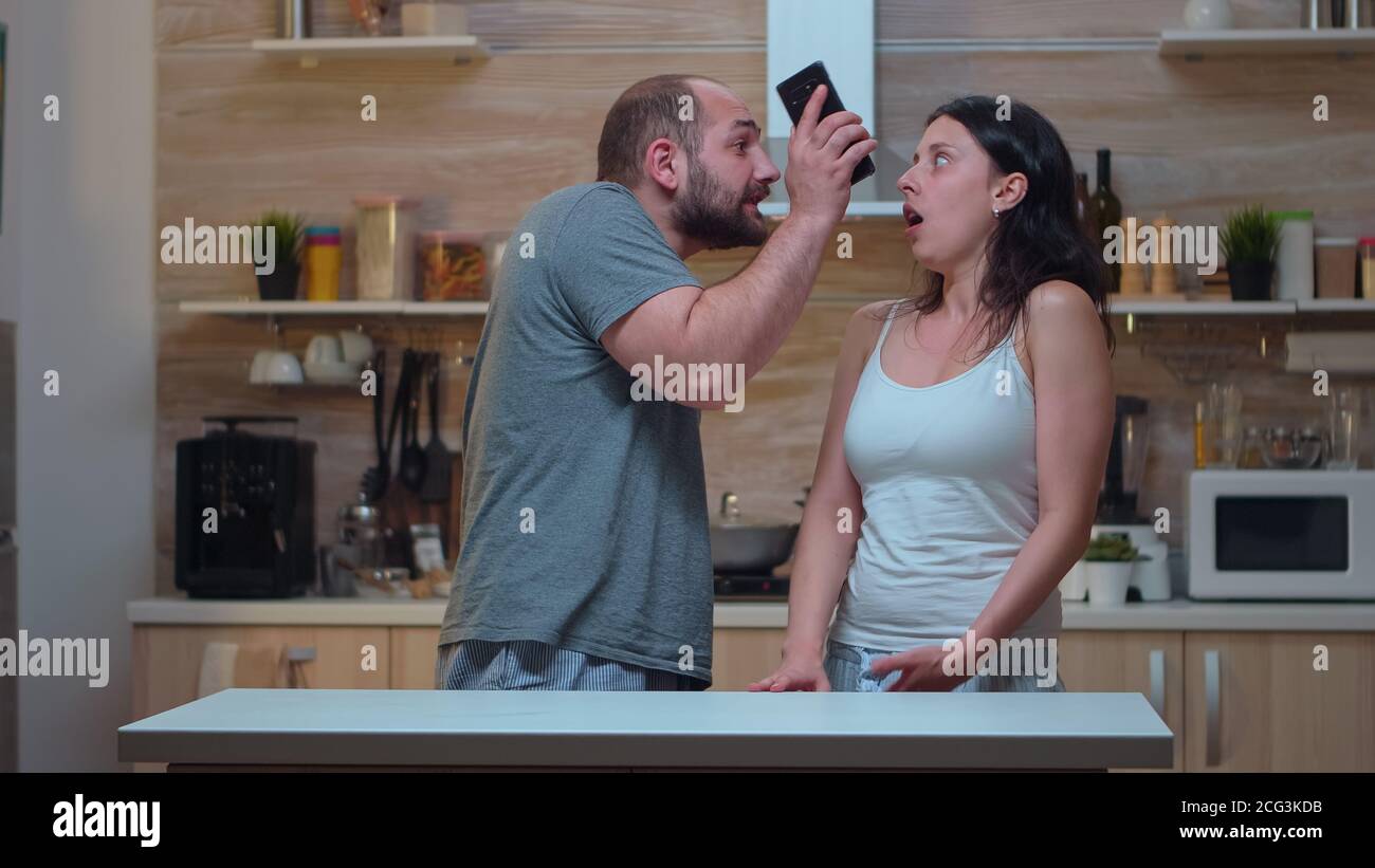 Angry man yelling at unfaithful wife in the kitchen. Jealous partner cheated frustrated offended irritated accusing woman of infidelity arguing her with messages on smartphone screaming desperate Stock Photo