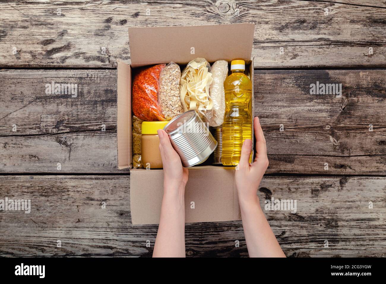 Female hands packing donation box with food items of staple products on wooden table. Person woman receiving donation Food box. Donate Food delivery Stock Photo