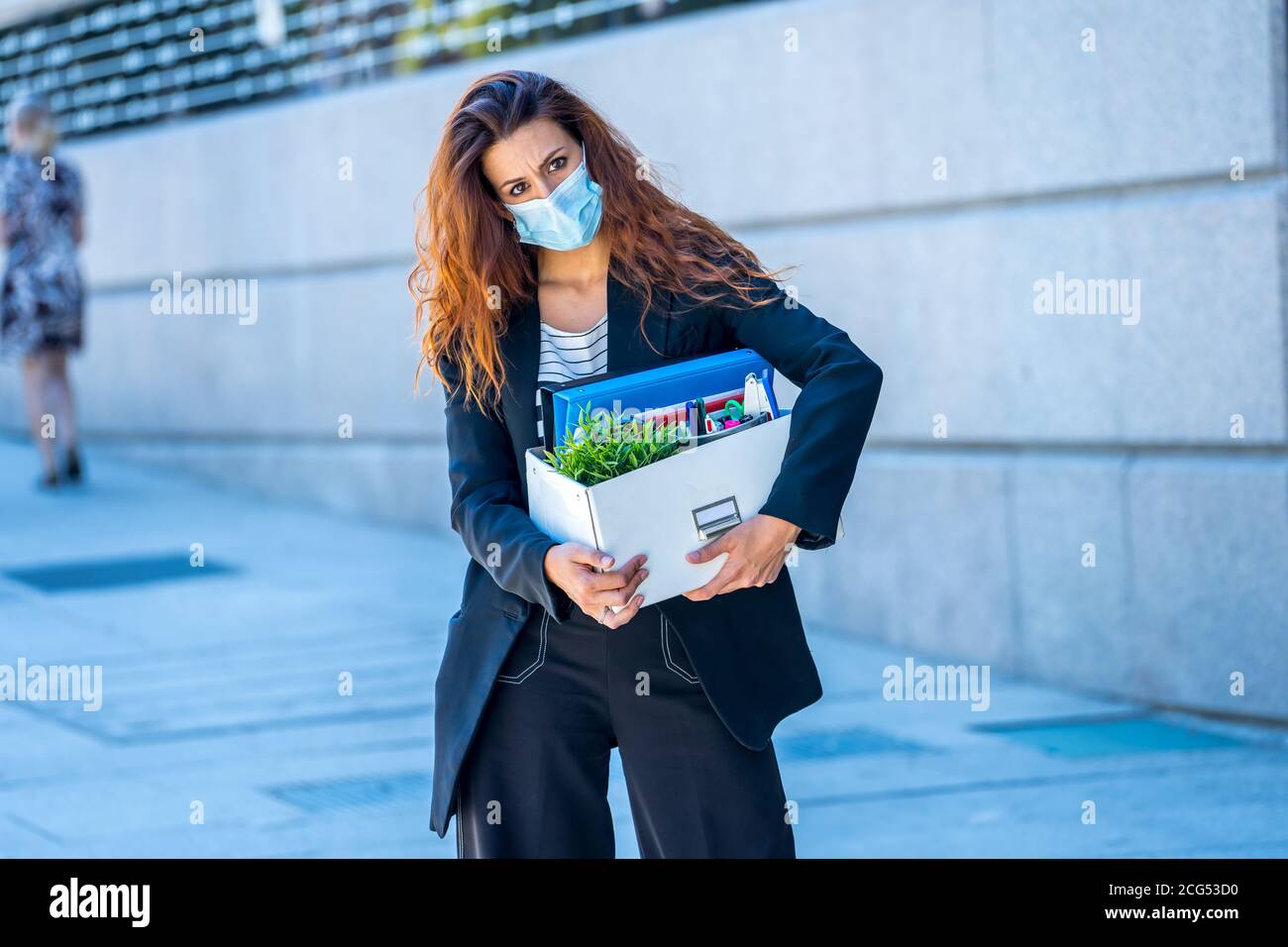 Depressed business woman with face mask outside office with personal staff box feeling hopeless after being fired from work. Coronavirus job losses, l Stock Photo