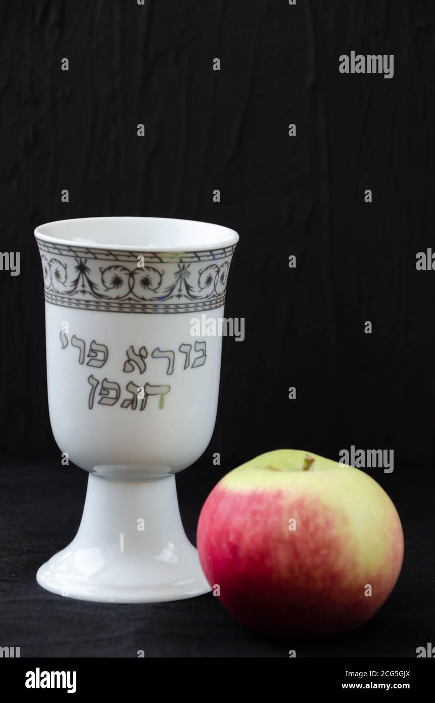 White porcelain kiddush wine cup and an apple on black background. The inscription on the cup in Hebrew: Creator of the fruit of the vine Stock Photo