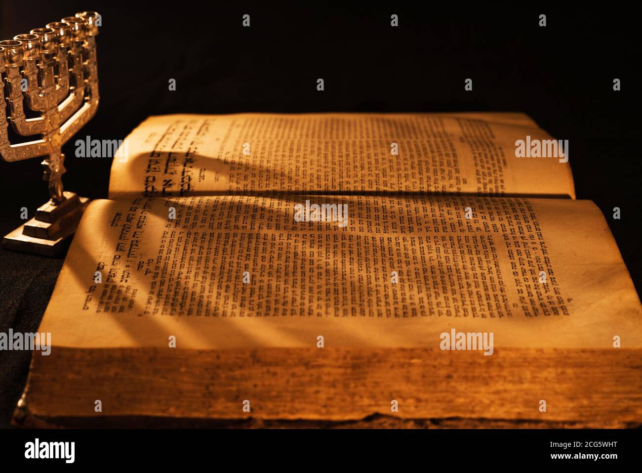 Hebrew Bible and silver menorah in light of burning candle on dark background. Shadow from menorah on open pages of Jewish prayer book in the darkness Stock Photo