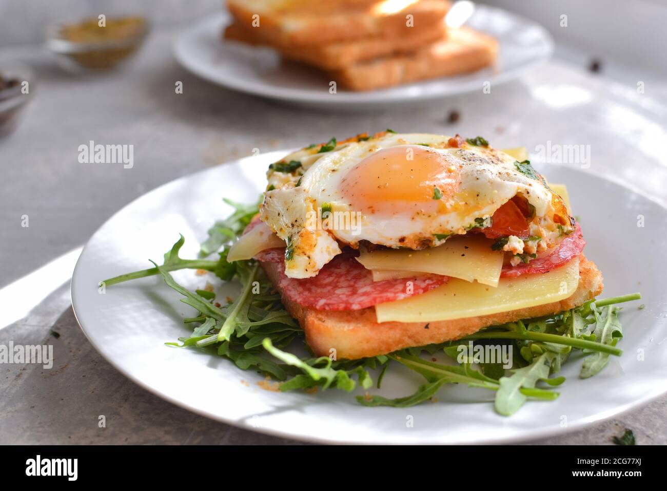 Tasty breakfast. Toast with egg, cheese and sausage. Sandwich in a white plate with arugula. Light background. Closeup Stock Photo