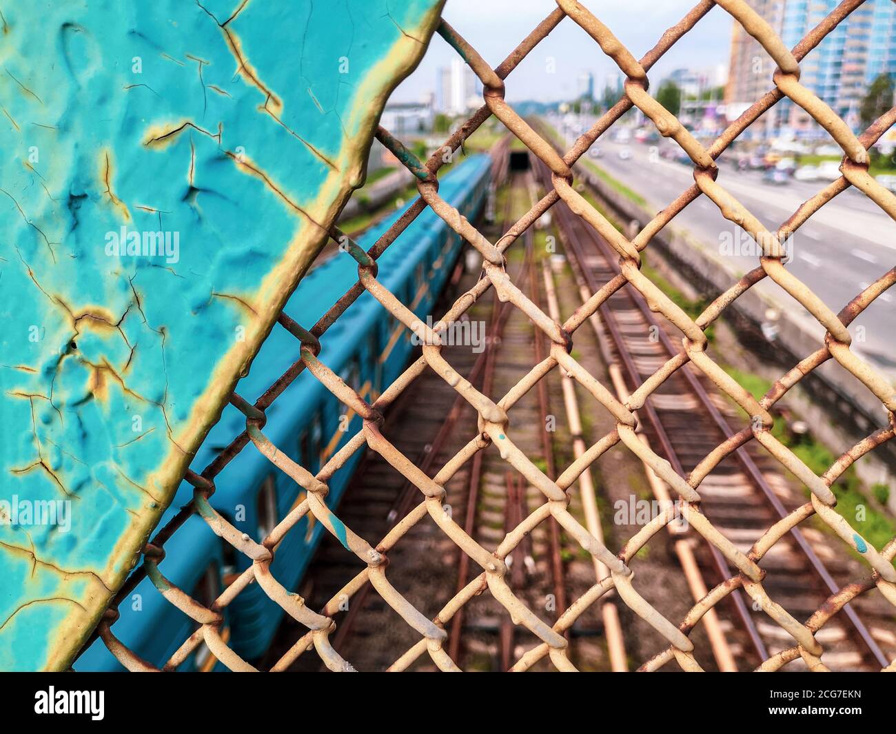 View through the old rusty painted cracked chainlink fence over the railway with blue tube train. Urban cityscape from the bridge. Stock Photo
