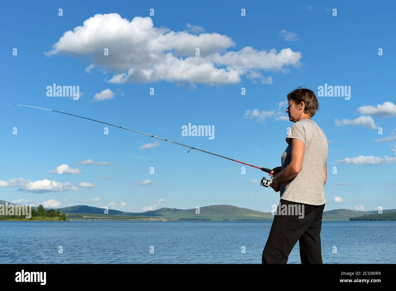 Woman fisherman catches a fish from a blue lake against a blue sky with clouds. Stock Photo