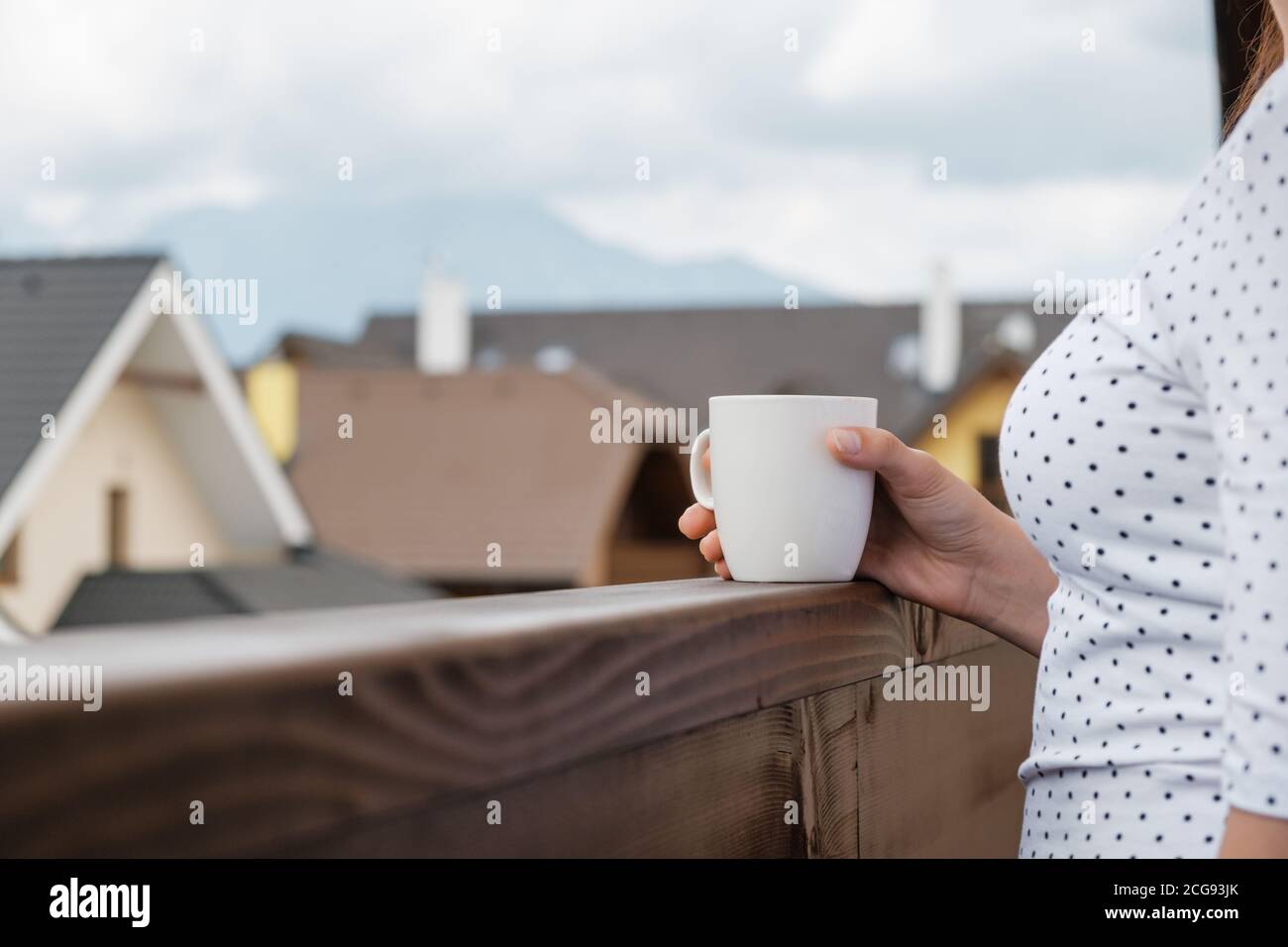 Close up a white cup in a woman hand on the wooden balcony on the background of mountains and house roof. Morning on the vacation Stock Photo