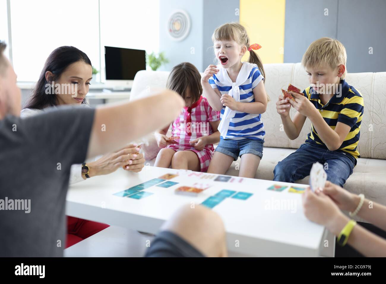 Adults and children are sitting at table and holding playing cards girl stood up and shouts. Stock Photo