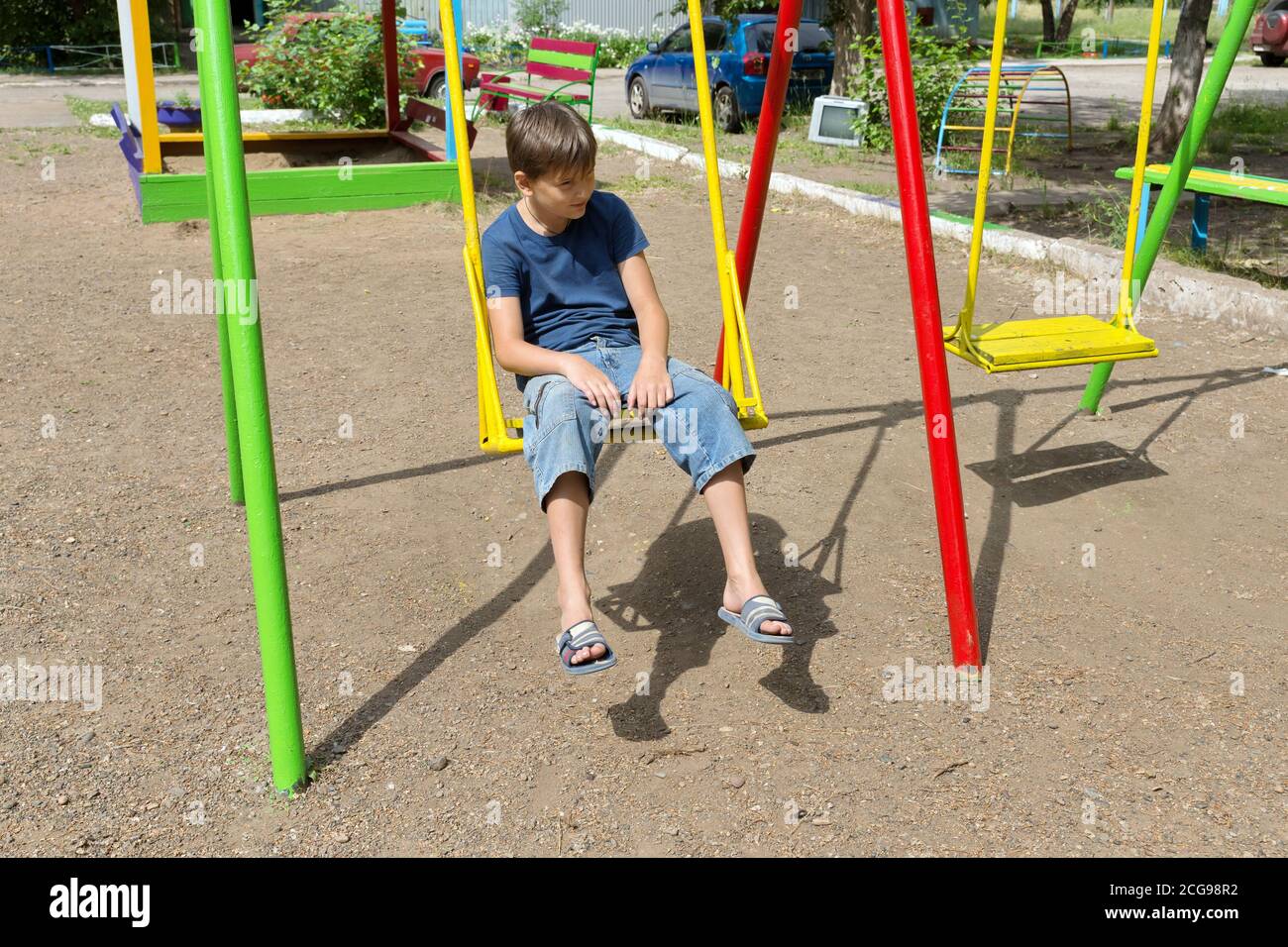 A bored boy sits on a swing in the courtyard of a residential building. Stock Photo