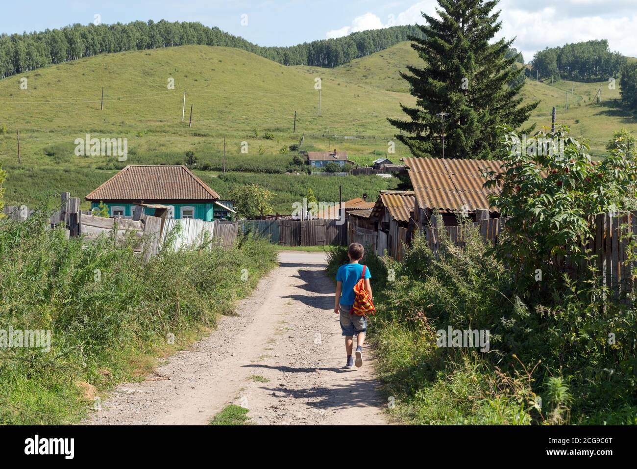 A boy, 12 years old, with a backpack is walking along a dirt road past a wooden fence in the village on a summer sunny day. Stock Photo