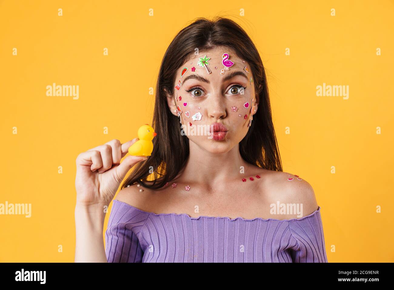 Image of young joyful woman with stickers on face holding rubber duck isolated over yellow background Stock Photo