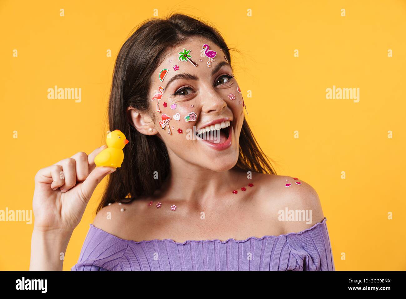 Image of young joyful woman with stickers on face holding rubber duck isolated over yellow background Stock Photo