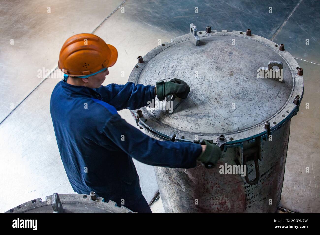 Worker in blue work wear and orange hardhat and grey metal scratched metallurgical bucket with metal titanium sponge. Raw material for clear titanium. Stock Photo