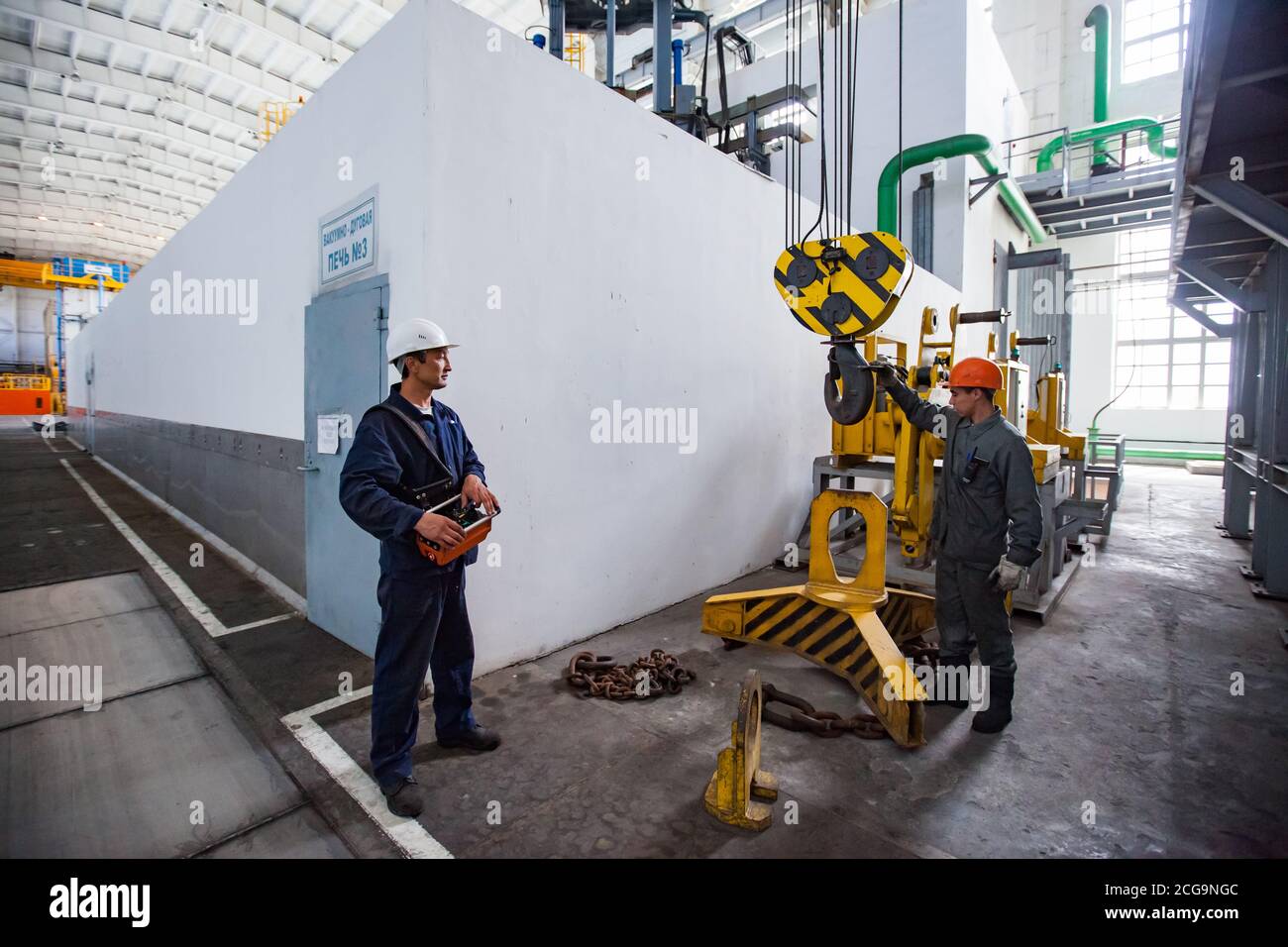 Workers using crane on titanium metallurgy plant. For loading titanium sponge to vacuum arc furnace. Furnace on foreground. Stock Photo
