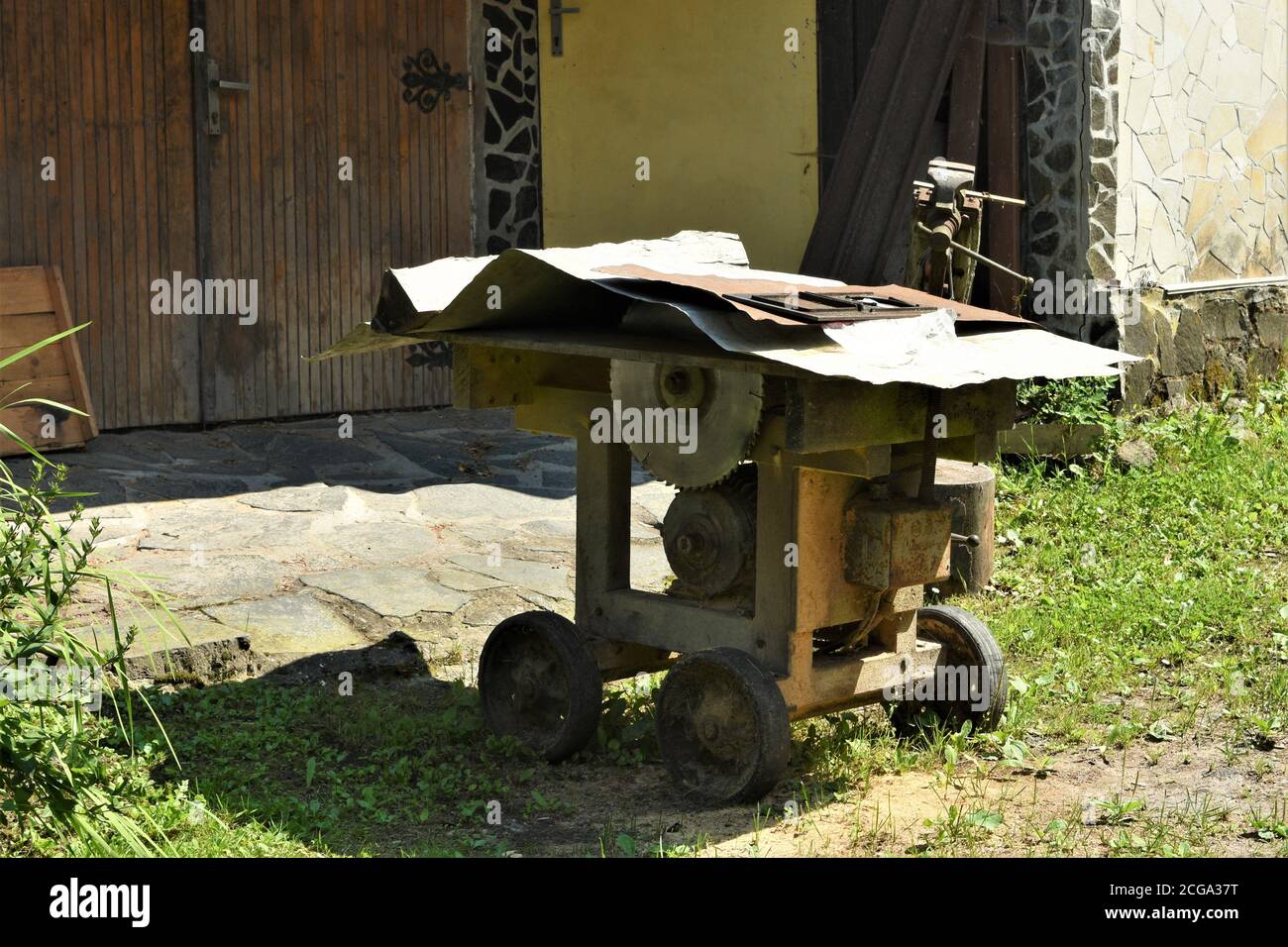 Old circular table saw machine or circular saw bench on four wheels covered with peaces of metal sheets standing on the grass in front of gate. Stock Photo