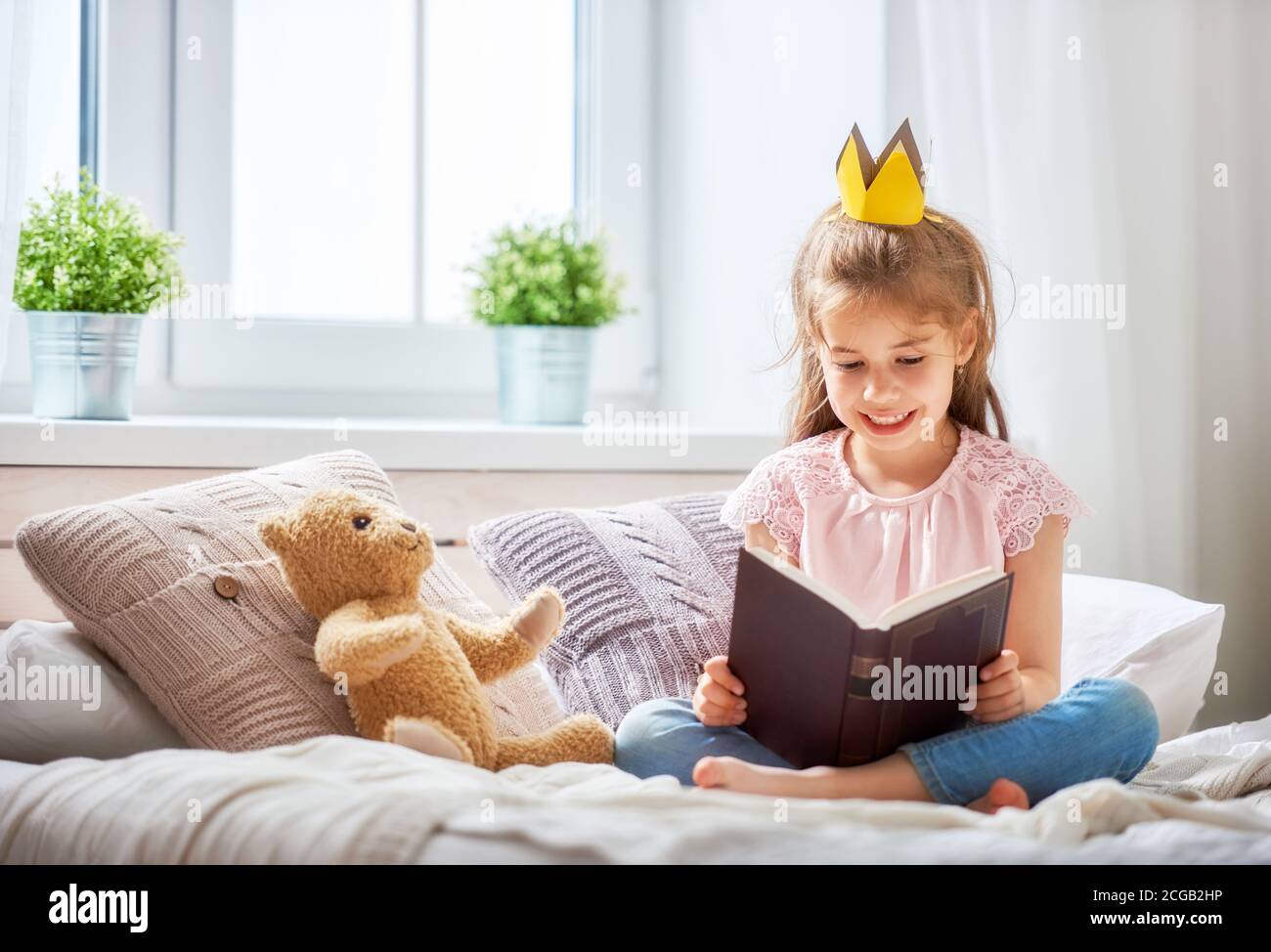 Cute little child girl reading a book in the bedroom. Kid with crown sitting on the bed near window. Stock Photo