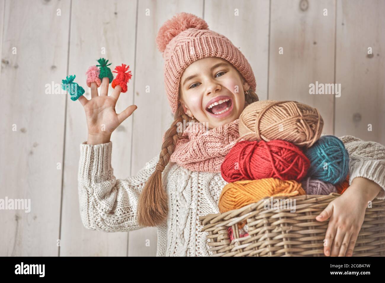 Winter portrait of happy little girl wearing knitted hat, scarf and sweater. Child holding a basket of skeins of yarn and having small hats on her fin Stock Photo