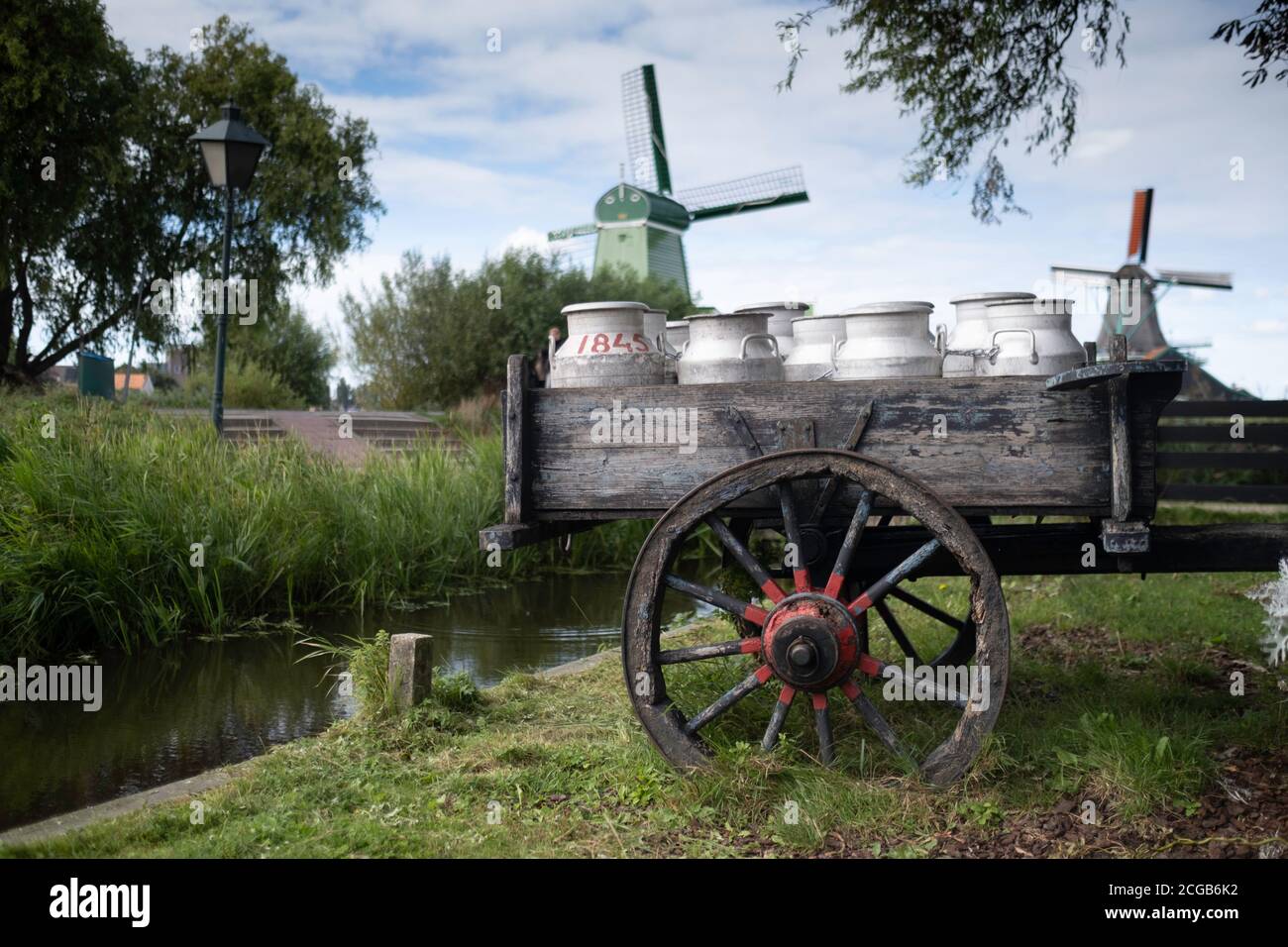 Milk cans on an old wheel cart in a garden in the Zaanse Schans in the Netherlands with windmills in background Stock Photo