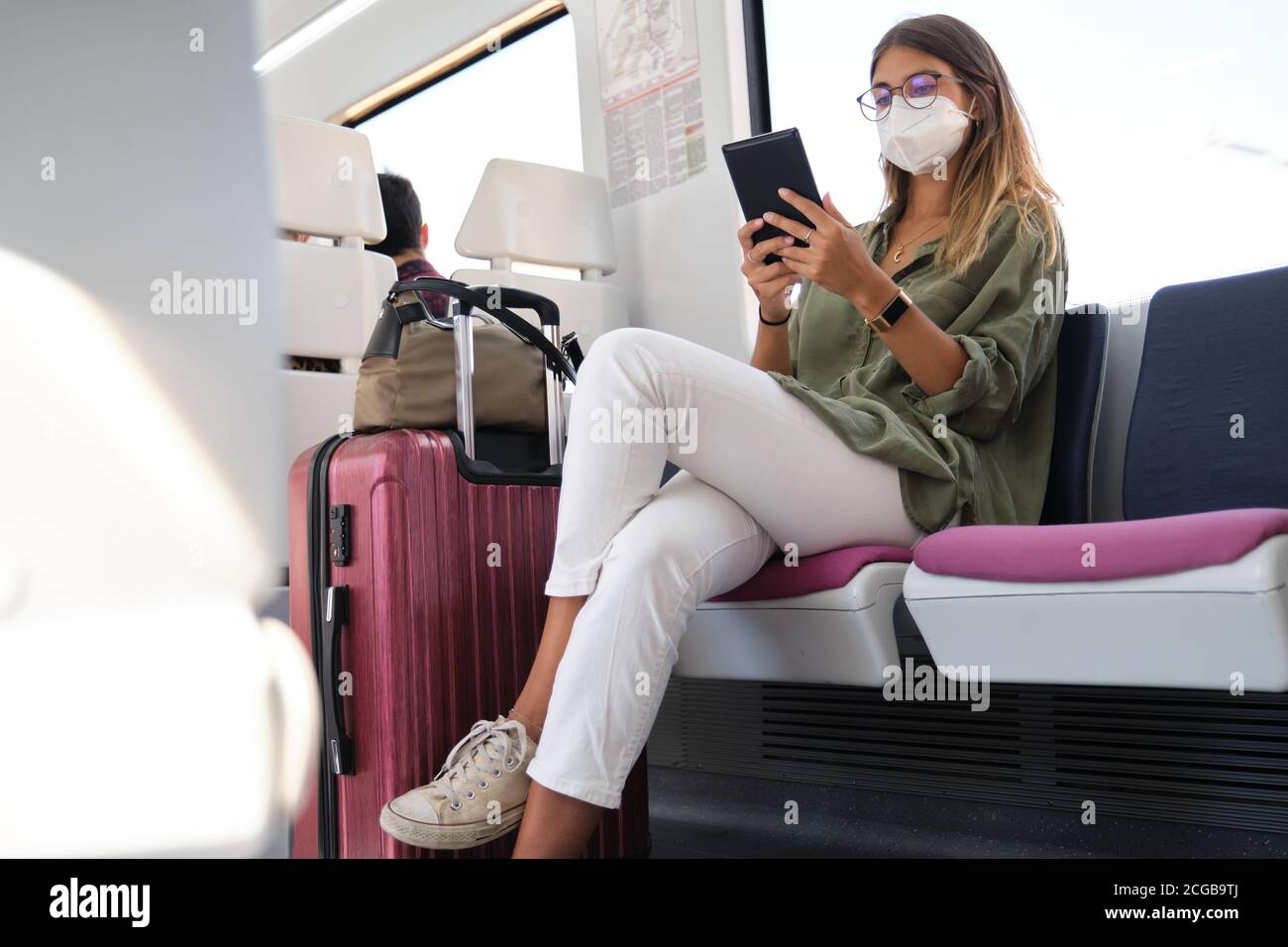 Young woman wearing face mask and carrying a suitcase reading a e-book in the train. New normal travel concept. Stock Photo