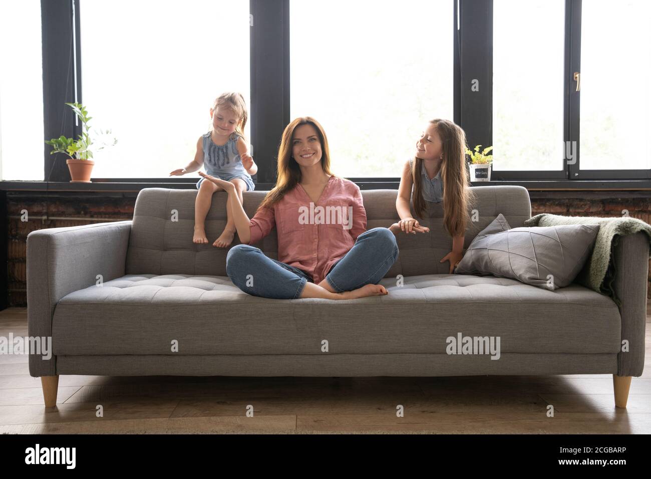 Calm woman concentrating on yoga exercises on couch at home while two noisy kids laughing, running Stock Photo