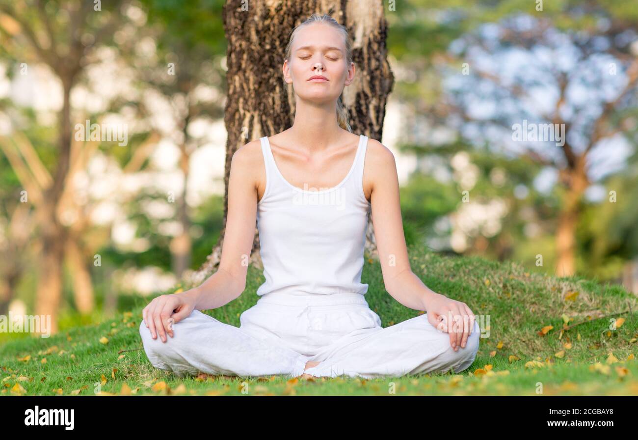 Calm young woman meditating outdoor on the grass at the park on a peaceful morning. Self care, mind and body relaxation. Stock Photo