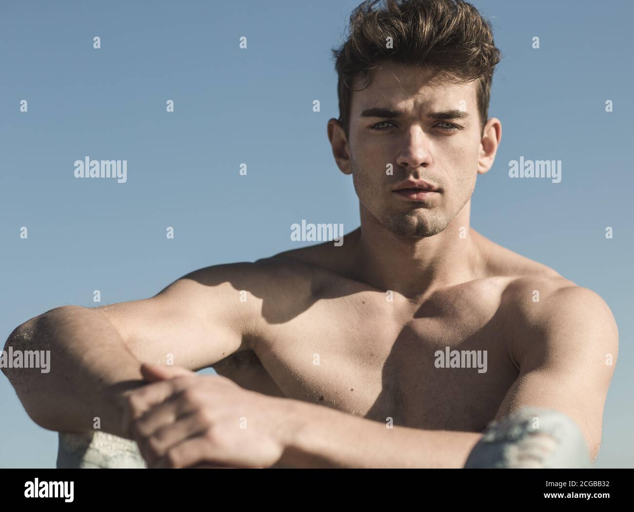 Sexy portrait of a handsome male model with beautiful eyes sitting at the beach and looking at the camera. Stock Photo