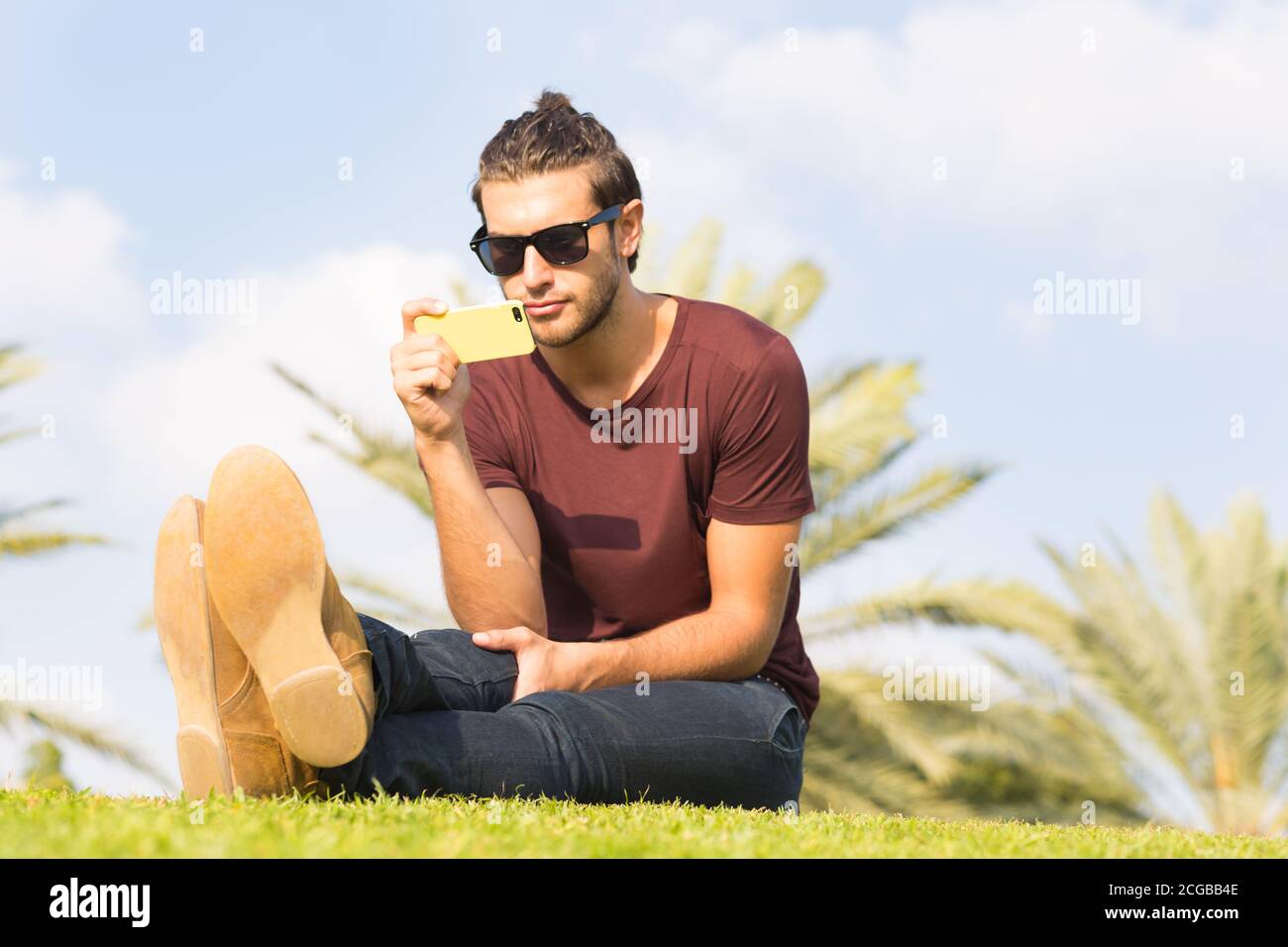 Handsome young man using a mobile phone while sitting on the grass in the park on a sunny summer day. Stock Photo