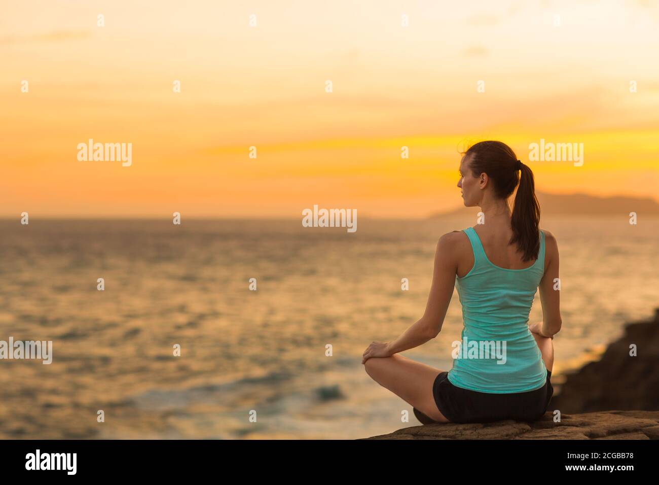 Fit relaxed woman meditating at peace on the beach with a beautiful view of the ocean during sunset. Health, yoga and harmony. Stock Photo