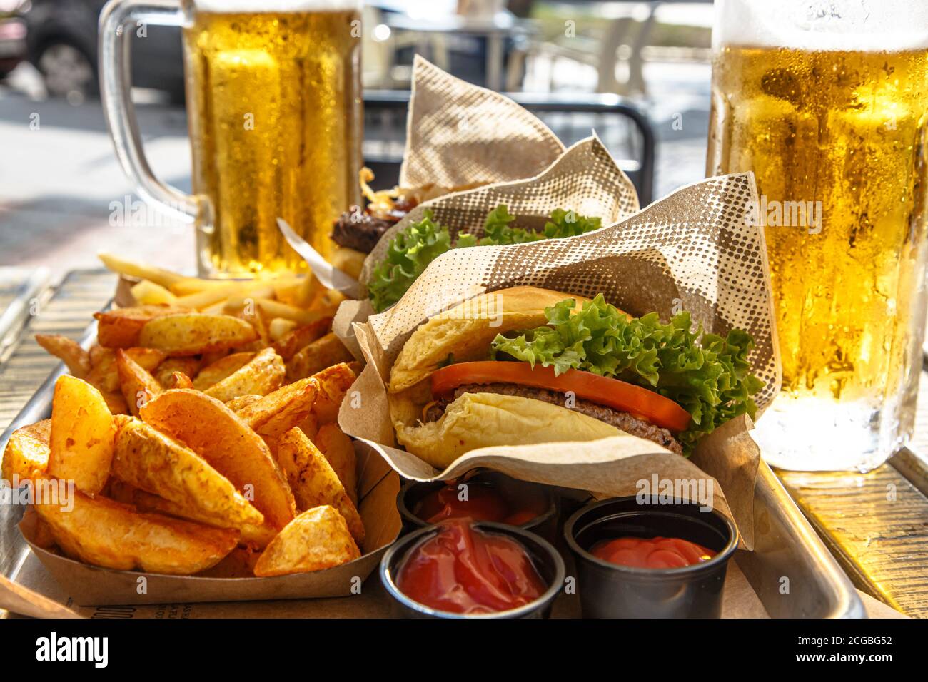 Burgers, potatoes, ketchup and mugs of beer on the table of a street cafe. Stock Photo