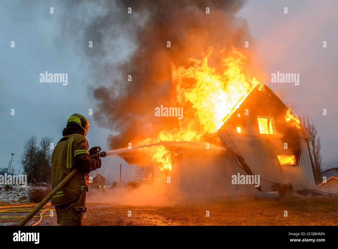 Norwegian first responder firefighter from behind trying to put out flames on house on fire with huge flames and smoke, in winter with snow. Stock Photo