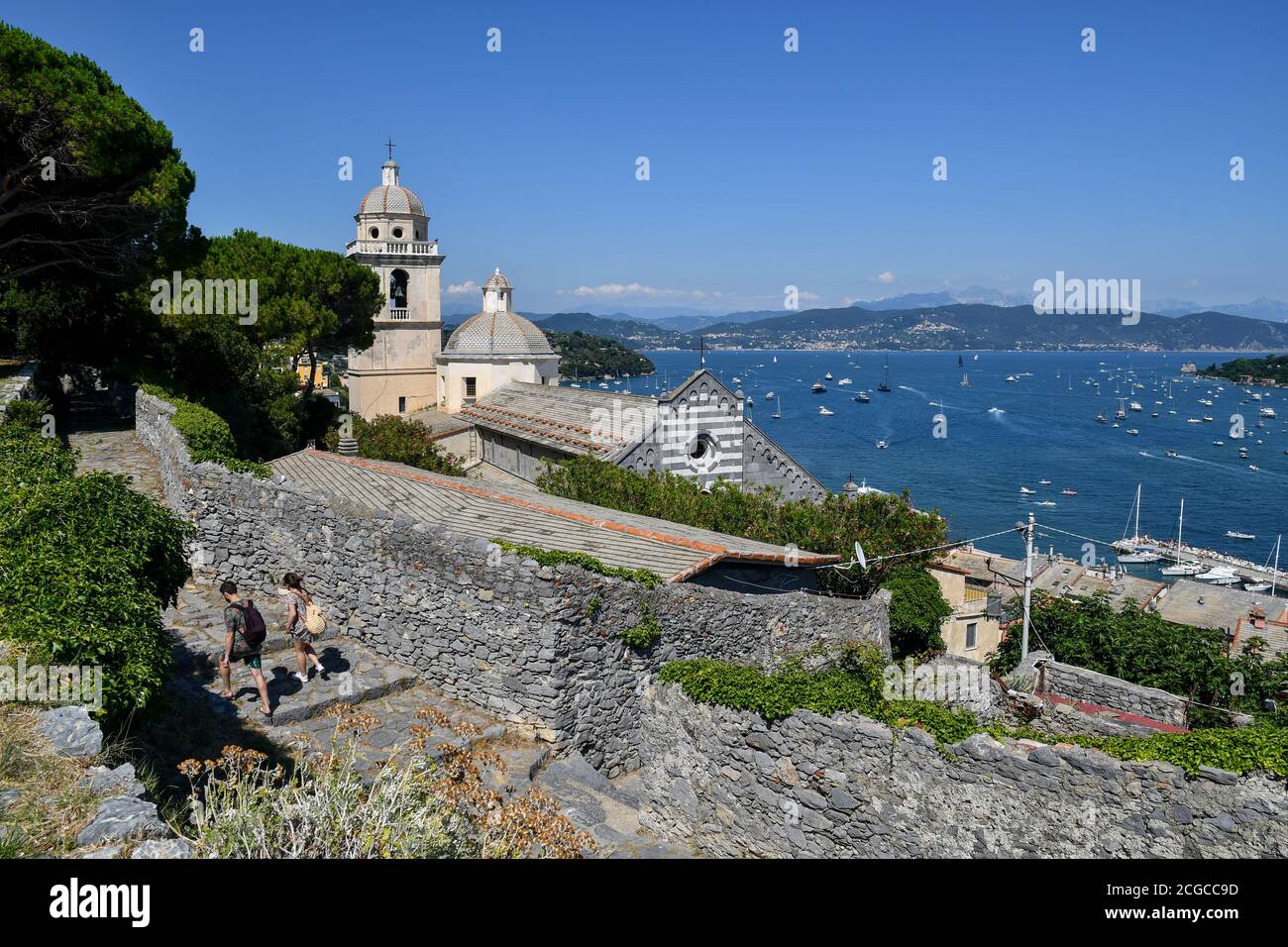Elevated view of the Gulf of Poets with the church of St Lawrence (12th century) and a couple of hikers in summer, Porto Venere, La Spezia, Italy Stock Photo