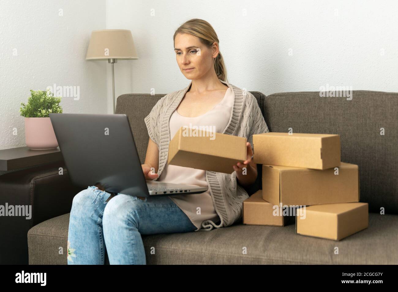 young woman small online business owner sitting on couch and preparing package for shipping on laptop at home. sme entrepreneur Stock Photo