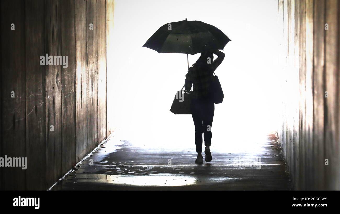 silhouette of a lady woman holding an umbrella emerging from a dark tunnel. Wet weather working city chic lifestyle.Female in the rain and puddles Stock Photo