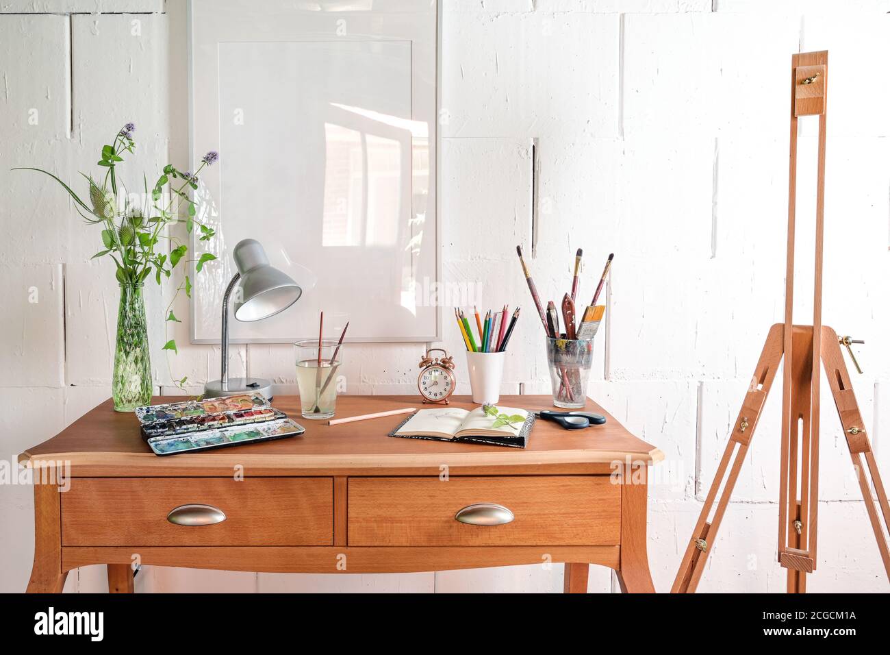 Creative place for art work or hobby, crayons, sketchbook, watercolor and brushes on a small wooden desk and an empty easel against a white painted wa Stock Photo