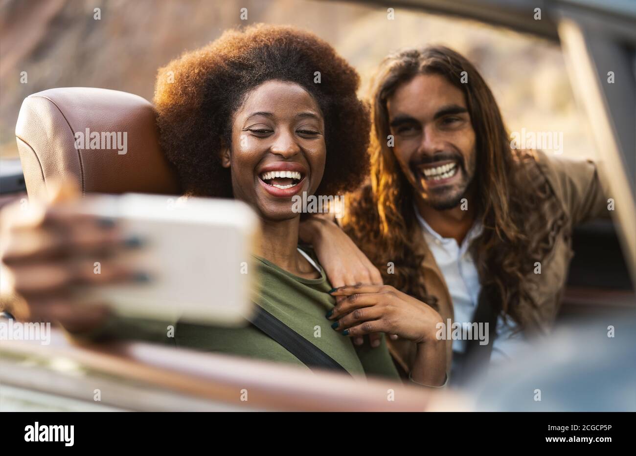 Happy young couple taking selfie with mobile smartphone while doing road trip - Travel people having fun driving in convertible car Stock Photo