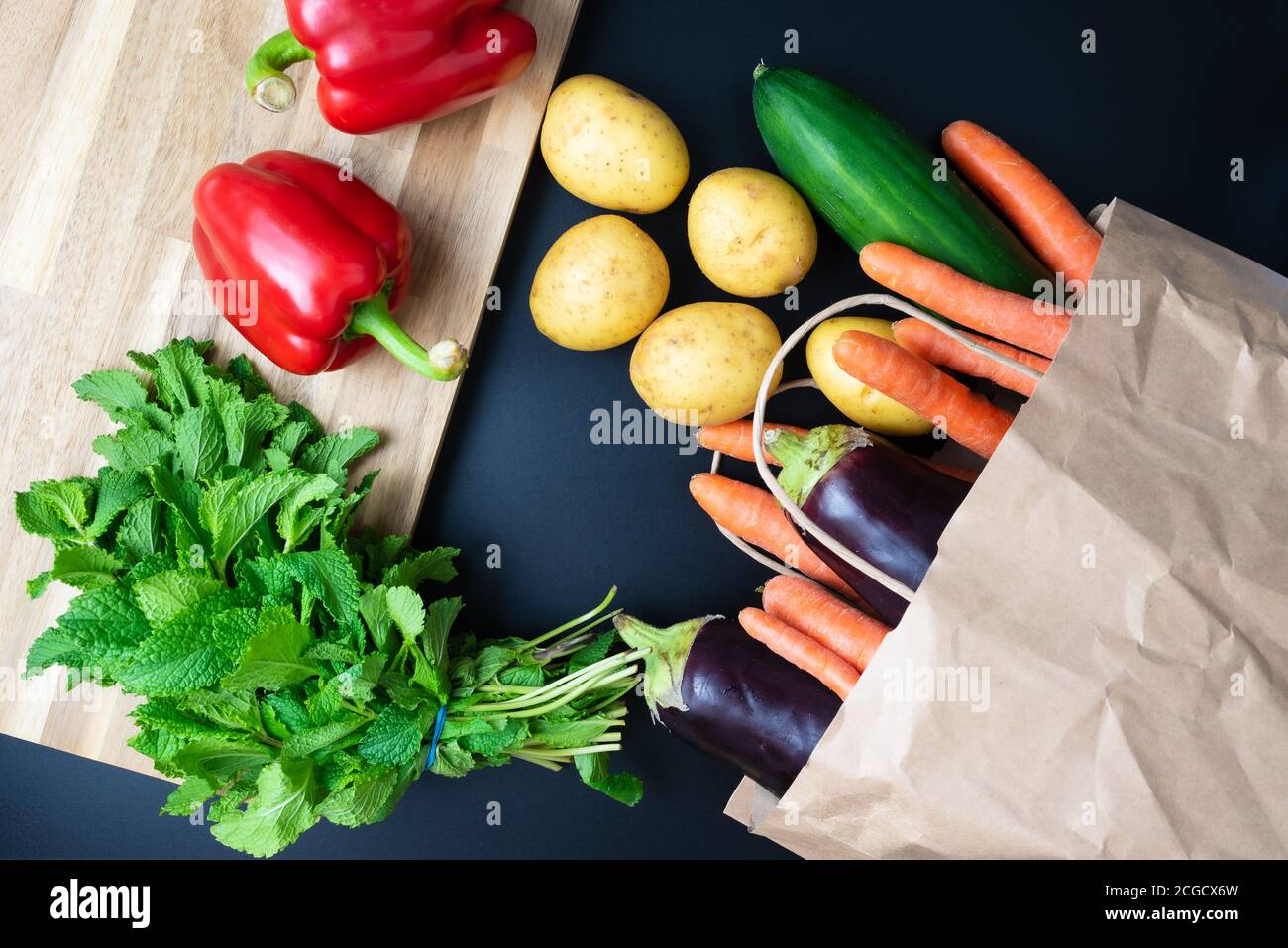 fresh organic vegetables spilling out of paper shopping bag on dark kitchen counter with wooden cutting board, healthy eating and cooking concept Stock Photo